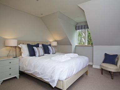 A bedroom with a bed with white bedding and blue pillows a nightstand with a lamp and a chair by a window at 3 The Drive Hillfield Village Dartmouth