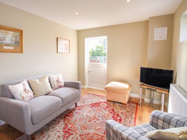 A living room with a gray sofa, a patterned armchair, a beige ottoman, a television on a glass stand, and wall art at Roundham Cottage in West Bay