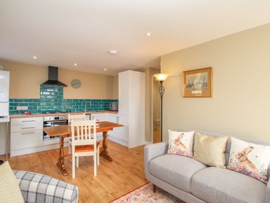 An open plan living room and kitchen with a wooden dining table and chairs and a gray sofa with animal print cushions at Roundham Cottage in West Bay