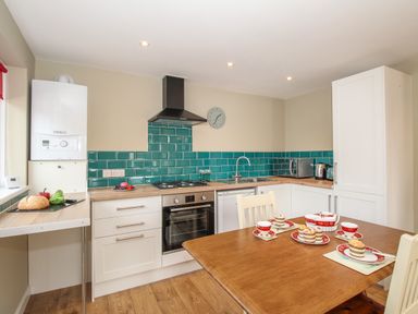 A kitchen with white cabinets teal tile backsplash a wooden table with teacups and cakes at Roundham Cottage in West Bay