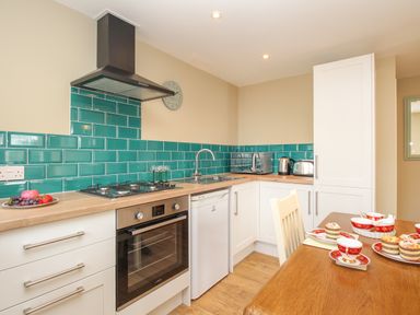 A kitchen with white cabinets green tiled backsplash wooden countertop and a dining table with cups and cakes at Roundham Cottage in West Bay