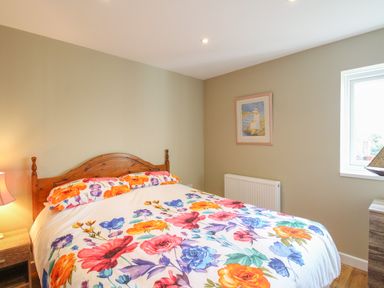 A bedroom with a wooden bed frame and floral bedding a bedside table with a lamp a radiator and a window at Roundham Cottage in West Bay