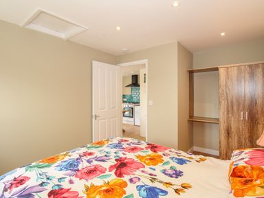 A bedroom with a floral bedspread a wooden wardrobe and a view through an open door to a kitchen at Roundham Cottage in West Bay