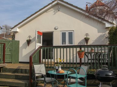 A patio area with a small round table set with bread and wine glasses chairs near a barbecue grill outside a white house at Roundham Cottage in West Bay