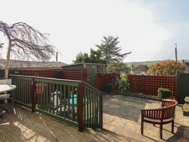 A backyard with wooden deck stairs leading to a patio with a wooden chair a round table and a small shed with lattice fencing at Roundham Cottage in West Bay