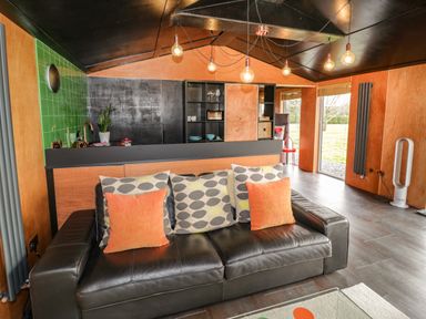 A living area with a black leather sofa with patterned and orange cushions a kitchen area with green tiles and shelving and large glass doors at The Tractor Shed in Lower Broadheath Worcester