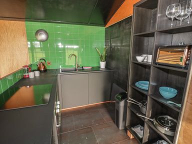 A kitchen with green tiled wall a black countertop a kettle a double sink a plant an open shelf with dishes pots and glasses at The Tractor Shed in Lower Broadheath Worcester