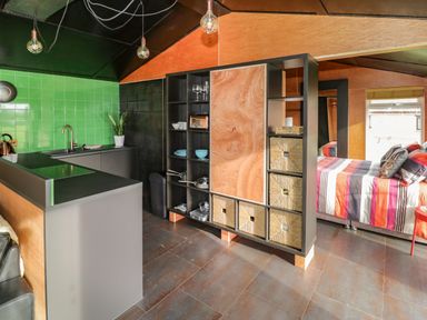 A kitchen with green tiled backsplash a black countertop a shelving unit and a bedroom with a striped bedspread at The Tractor Shed in Lower Broadheath Worcester