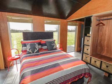 A bedroom with a bed with striped bedding and pillows near windows and a shelving unit with storage baskets at The Tractor Shed in Lower Broadheath Worcester