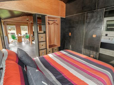 A bedroom with a striped bedspread a wardrobe with storage compartments and a view into a dining area with green chairs at The Tractor Shed in Lower Broadheath Worcester