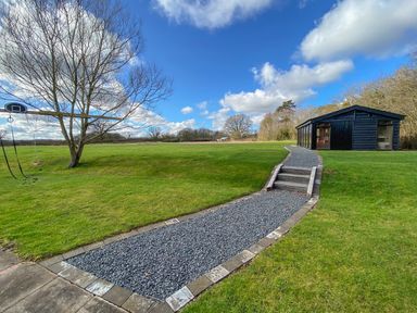 An outdoor grassy area with a paved and gravel path leading to a black building and a tree with a basketball hoop and swings at The Tractor Shed in Lower Broadheath Worcester