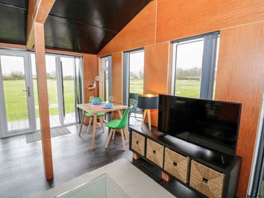 A dining table with green chairs and tableware beside a TV on a black stand with baskets in a room with large windows at The Tractor Shed in Lower Broadheath Worcester