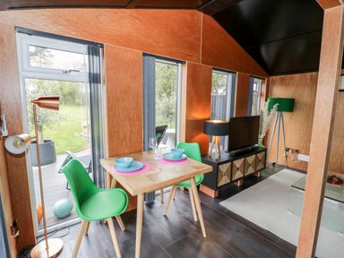 A dining area with a wooden table and green chairs next to windows and a living space with a television on a black stand at The Tractor Shed in Lower Broadheath Worcester