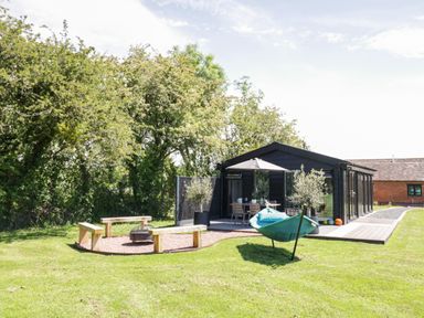 A backyard with wooden benches around a fire pit a green hammock and a black shed with a patio at The Tractor Shed in Lower Broadheath Worcester