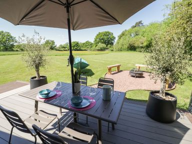 An outdoor dining table with chairs and an umbrella on a deck overlooking a lawn with a fire pit and wooden benches at The Tractor Shed in Lower Broadheath Worcester