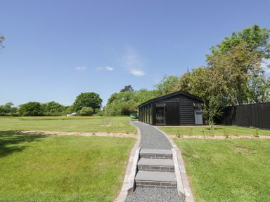 A gravel path with steps leading to a black shed in a green garden with trees and a blue sky at The Tractor Shed in Lower Broadheath Worcester