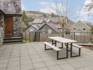 A patio area with a picnic table and benches near stone buildings and a mountain in the background at Bryn Elsi in Betws-Y-Coed