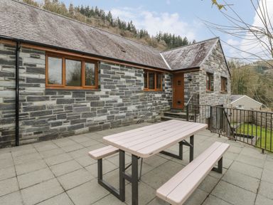 An outdoor patio with a picnic table and stone house wall with windows at Bryn Elsi in Betws Y Coed