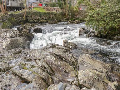 A flowing rocky river with some greenery and stone walls and buildings in the background at Bryn Elsi in Betws-Y-Coed