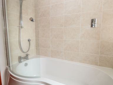 A bathroom with a white bathtub beige tiled walls a silver shower head and a curved glass shower screen at Seaview Cottage in Salthouse Norfolk