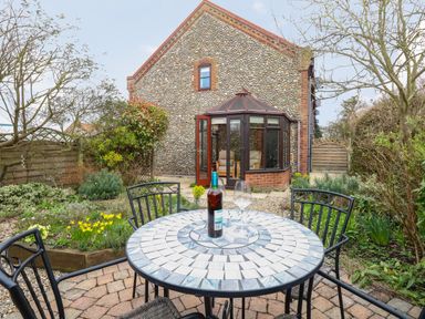A garden patio with a round mosaic table and chairs with a bottle and glass in front of a conservatory at Seaview Cottage in Salthouse Norfolk