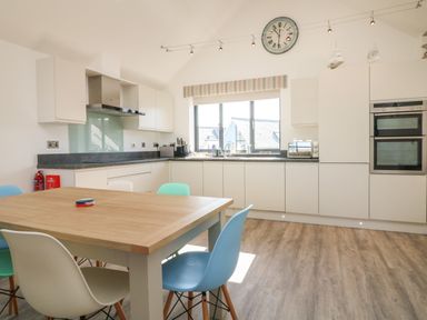 A kitchen with a wooden dining table and colored chairs a clock above a window and built in appliances at Seaquel in Westward Ho