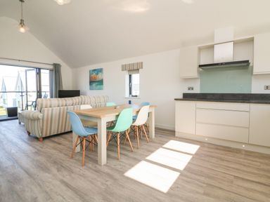 An open-plan room with a dining table and chairs a striped sofa kitchen cabinets and a balcony door at Seaquel in Westward Ho
