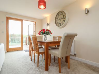 A dining room with a wooden table and six chairs a large wall clock and glass doors at Maple in Falmouth