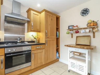 A kitchen with wooden cabinets an oven a stovetop a white kitchen cart with a wooden top and a wall shelf with hanging mugs and fruit at Maple in Falmouth