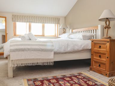 A bedroom with a bed with white linens and rolled towels a wooden nightstand with a lamp and a colorful patterned rug at Maple in Falmouth