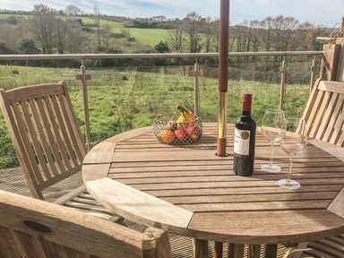 A wooden outdoor table with a bottle of wine two wine glasses and a basket of fruit on a balcony with green fields and trees in the background at Maple in Falmouth