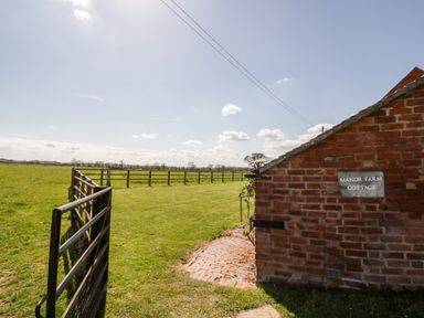 A metal gate opening to a grassy field beside a brick wall with a Manor Farm Cottage sign at Manor Farm Cottage in Hill Croome near Upton Upon Severn