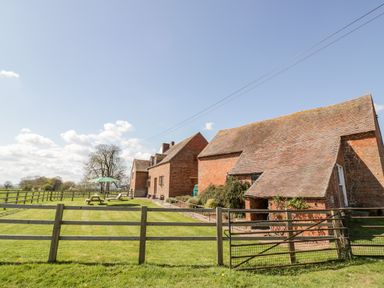 A fenced grassy yard with picnic tables and an umbrella next to brick farm buildings at Manor Farm Cottage in Hill Croome near Upton Upon Severn