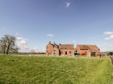 A grassy field with a wooden fence and brick farm buildings under a blue sky at Manor Farm Cottage in Hill Croome near Upton Upon Severn