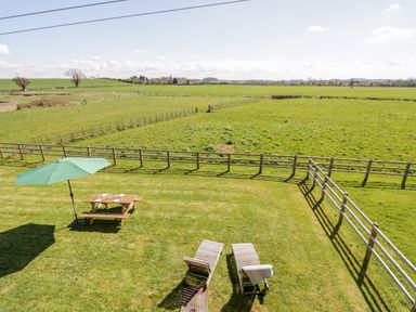 A grassy fenced backyard with two wooden lounge chairs and a picnic table with a green umbrella and plates at Manor Farm Cottage in Hill Croome near Upton Upon Severn