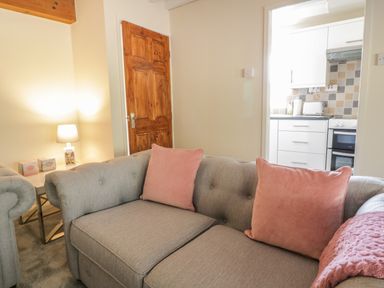 A living room with a grey sofa with pink cushions a wooden door a lamp and a view into a kitchen at The Old Smithy Cottage in Maentwrog near Penrhyndeudraeth