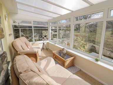 A conservatory with beige cushioned wicker chairs a wicker glass coffee table a telescope and large windows at The Old Smithy Cottage in Maentwrog near Penrhyndeudraeth