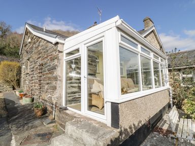An exterior view of a stone cottage with a white framed conservatory and outdoor seating at The Old Smithy Cottage in Maentwrog near Penrhyndeudraeth