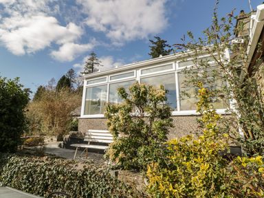 A garden with shrubs and a white bench outside a house with large windows at The Old Smithy Cottage in Maentwrog near Penrhyndeudraeth