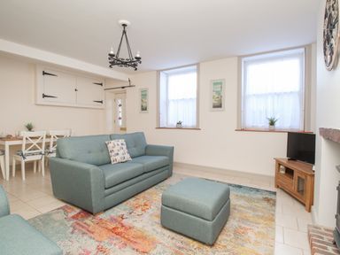 A living room with a blue sofa and ottoman a wooden TV stand with a TV two windows and a dining table with chairs at Bliss On The Bay in Weymouth