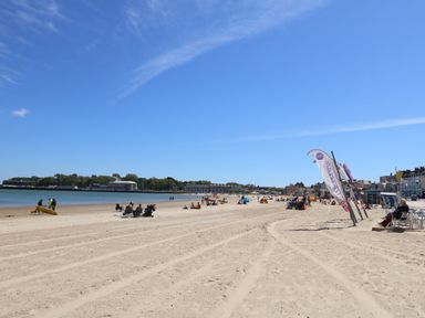 A sandy beach with people sitting and walking near the water and buildings in the background at Bliss On The Bay in Weymouth