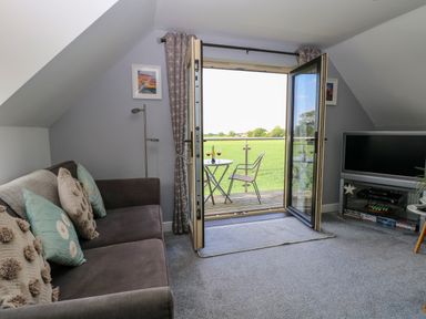 A living room with a sofa pillows and a TV near open double doors leading to a balcony with a table and chairs overlooking a field at Spinneys Den in Knapton