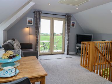 A sitting area with a sofa, floor lamp, television, and table with refreshments next to glass doors leading to outdoor seating at Spinney's Den in Knapton