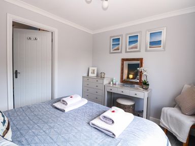 A bedroom with a double bed covered with a blue patterned blanket and white towels on top a grey chest of drawers and a dressing table with a mirror and a stool next to a white chair with cushions and a blanket on it three framed pictures hang on the wall above the dressing table Spinney's Den Knapton