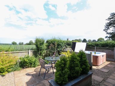 A garden patio with a round glass table and chairs next to a hot tub with a fence and greenery in the background at Spinney's Den in Knapton
