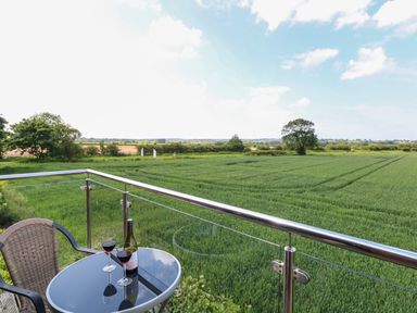 A balcony with a glass table holding a bottle of wine and two glasses overlooking a green field at Spinneys Den in Knapton