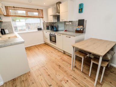 A kitchen with a wooden floor white cabinets a wooden table with stools and a window with a bamboo blind at Sea Snug in Spittal