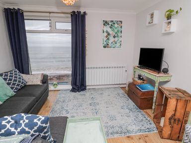 A living room with a sofa and cushions a television on a table and a large window with curtains showing a sea view at Sea Snug in Spittal
