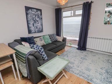 A living room with a dark sofa with patterned cushions a small table a rug and a large window with a sea view at Sea Snug in Spittal