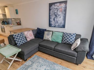 A gray sectional sofa with patterned cushions in a living room with wooden floor and a kitchen in the background at Sea Snug in Spittal